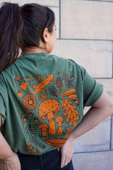 A picture of the back of the Forest Finds T shirt Print showing some berries, a salamander, a snail, a flower, a fern, some various types of mushrooms, some acorns, a pinecone branch, and some leaves.