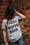 Kolbi lloyd wearing the TREES COFFE BEER T-Shirt with a little pine tree by "BEER" while standing in front of trees that are changing color during the fall. 