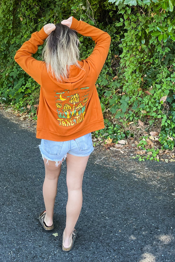 Kolbi wearing an orange zip hoodie with a mushroom design on the back that says Take a Trip at the bottom