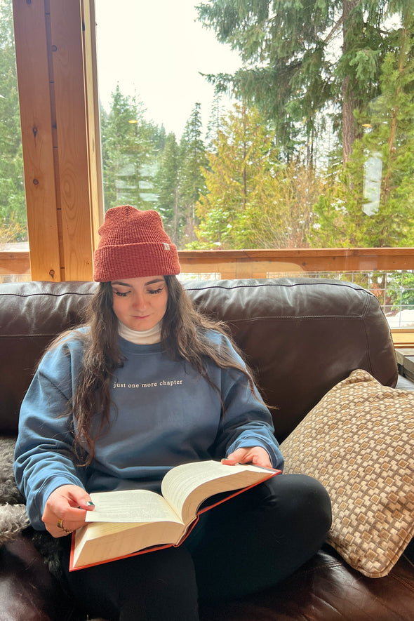 Person reading a book on a couch with a window view of trees wearing a sweatshirt that says "Just one more chapter"