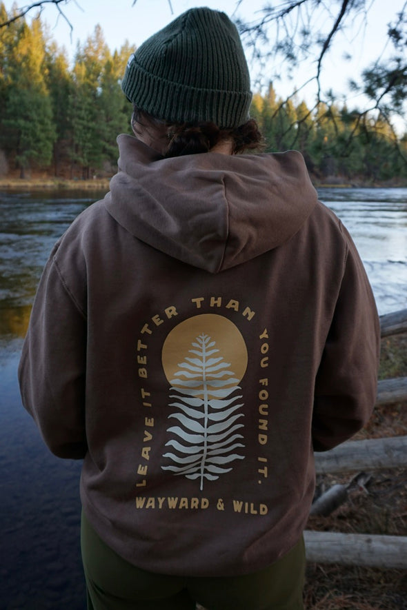 Person wearing a brown hoodie with a nature-themed design by a body of water.