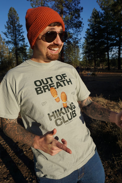 Man wearing a t-shirt with 'Out of Breath Hiking Club' text outdoors.