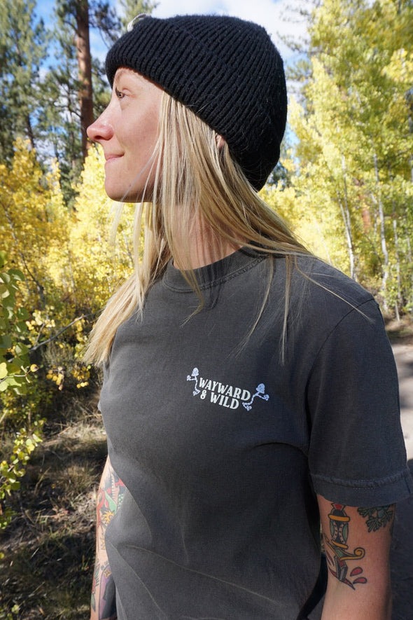 Person wearing a black beanie and t-shirt with 'Wayward & Wild' logo in a forest setting