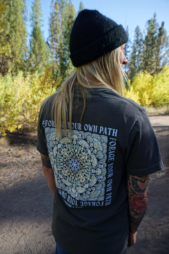 Person wearing a black t-shirt with a design and text, standing outdoors with trees in the background