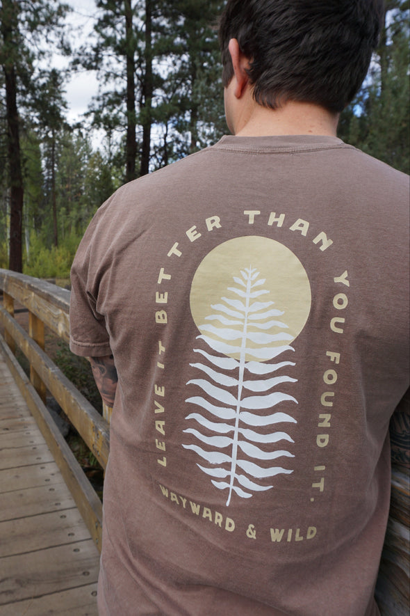Person wearing a brown t-shirt with a leaf design and text in a forest setting