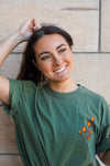 A photo of Kolbi looking off to the side with a hand on her head and showing the left chest screen print of the T shirt that shows a small cluster of mushrooms