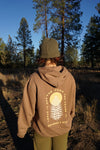 Person wearing a brown hoodie with a logo and text in a forest setting