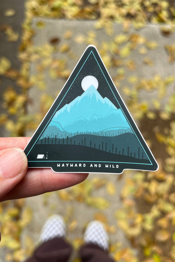 Triangular sticker with mountain design held by a hand on a blurred outdoor background