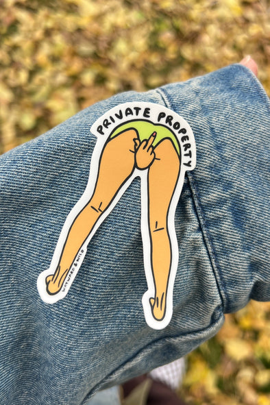 Sticker of legs with 'Private Property' text on a denim jacket against a blurred natural background