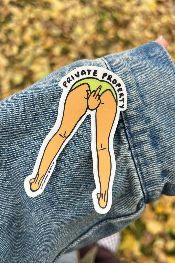 Sticker of legs with 'Private Property' text on a denim jacket against a blurred natural background