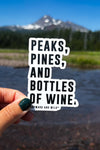 A up close photo of the peaks pines and bottles of wine sticker held up in front of a mountain and river landscape