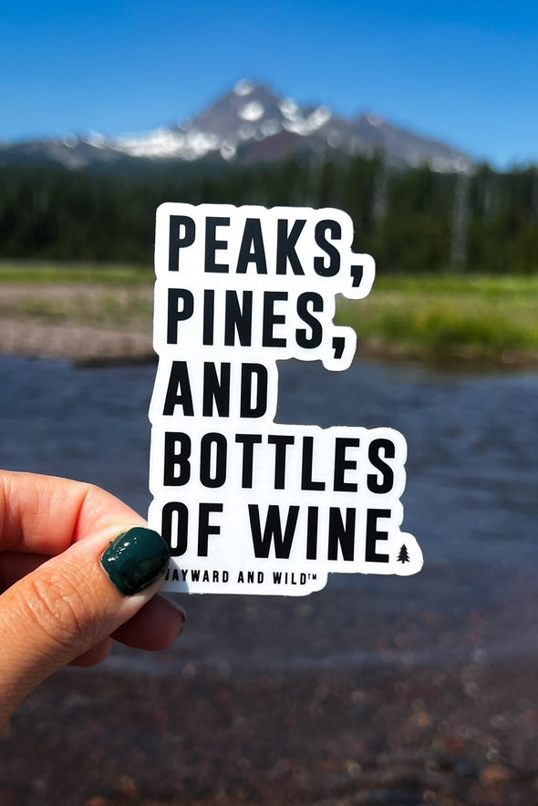 A up close photo of the peaks pines and bottles of wine sticker held up in front of a mountain and river landscape