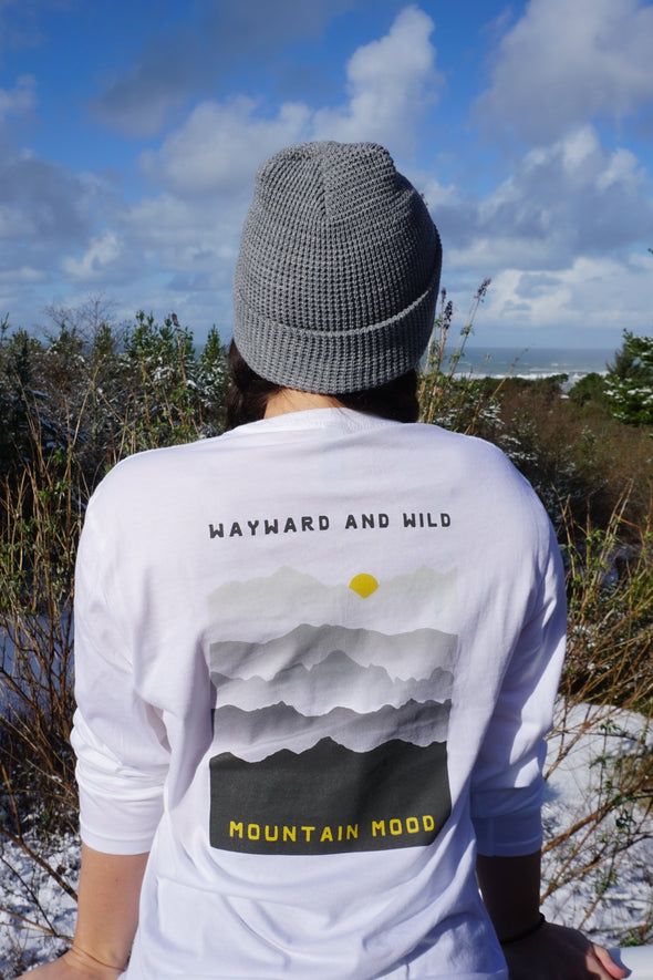  Kolbi leaning on the edge of a rail looking out at the distance while Wearing a white long sleeved T with the words Wayward & Wild and Mountain mood on a Grey mountain scape design printed on the back of the shirt