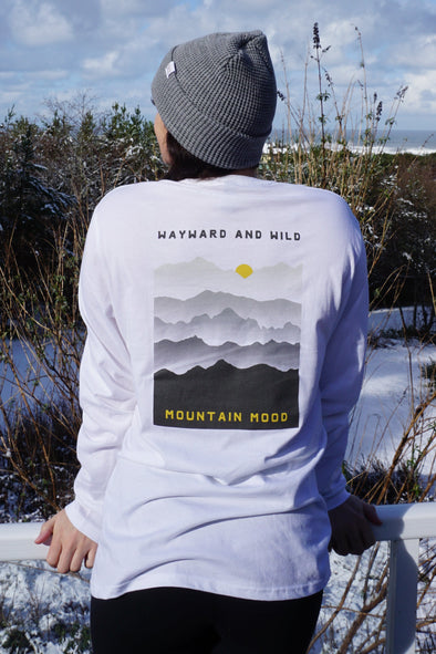 Kolbi Wearing a white long sleeved T with the words Wayward & Wild and Mountain mood on a Grey mountain scape design printed on the back of the shirt