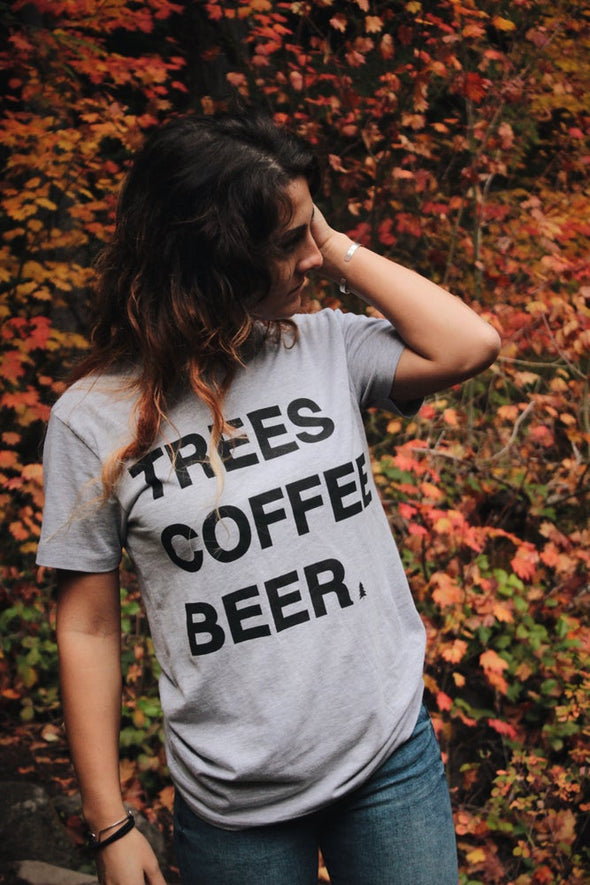 Kolbi lloyd wearing the TREES COFFE BEER T-Shirt with a little pine tree by "BEER" while standing in front of trees that are changing color during the fall. 