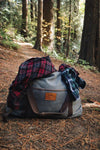 Picture of the gray Canvas Duffel Bag with a Wayward & Wild leather patch on the front sitting in a trail in the forest in Oregon. Duffel is styled with flannels coming out of the bag.