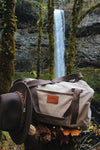Picture of the tan Canvas Duffel Bag with a Wayward & Wild leather patch on the front sitting in front of a waterfall in Silver Falls State Park in Oregon. Duffel is styled with a wool felt hat