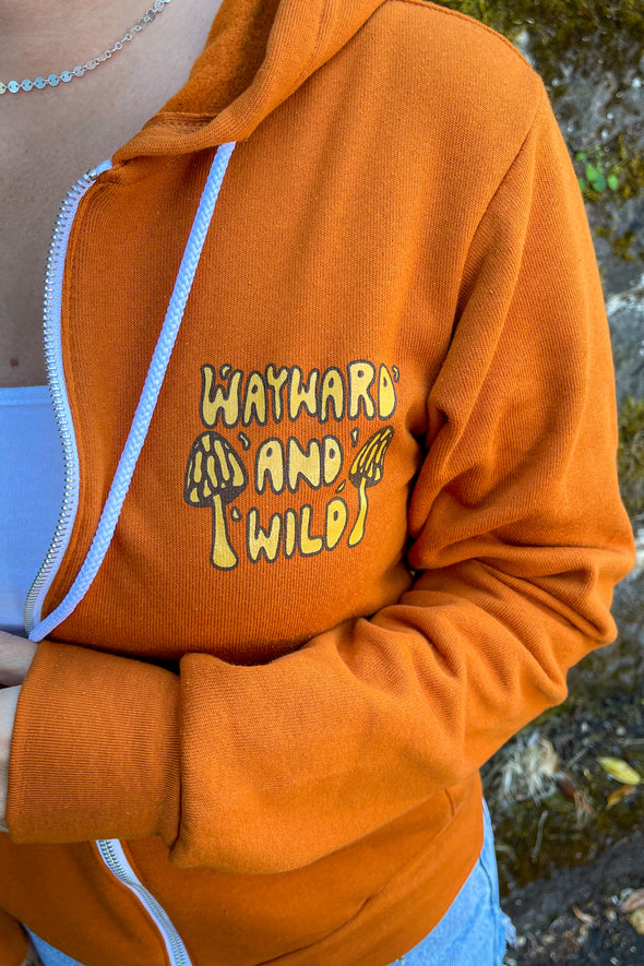 An up close shot of the left chest of the Orange zip "Take a trip" hoodie that says Wayward & Wild with two small mushrooms on each side