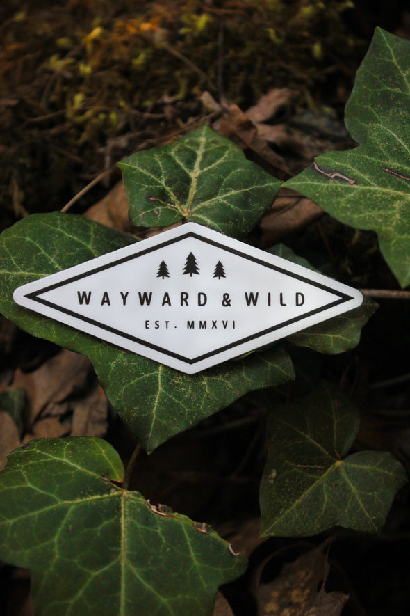 Wayward & Wild 3.5" Logo Sticker of the diamond logo that has three pine trees with "Wayward & Wild" below the trees and "EST. MMXV" below the Wayward & Wild. Sticker is laid out of some ivy leaves.