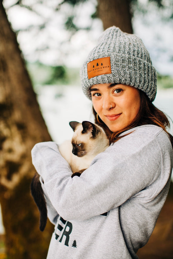 Photo of Stacie wearing the light gray Chunky knit Beanie with a leather patch the has three little pine trees "Wayward & Wild" below the trees and "EST. MMXVI" below that. 
