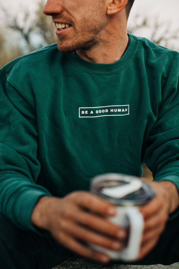 Picture of close view of the front of the pigment alpine green Be a Good Human crewneck sweatshirt on a guy holding a travel coffee cup