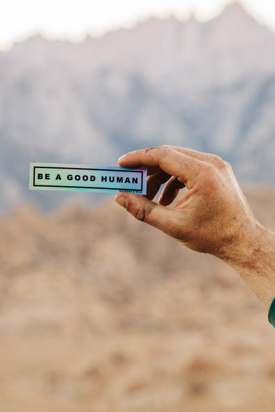 Photo of a guy holding the rectangular holographic sticker that says "Be A Good Human"