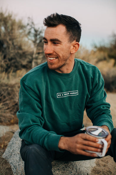 Guy smiling and holding a coffee cup while wearing the alpine pigment green Be a Good Human crewneck sweatshirt 